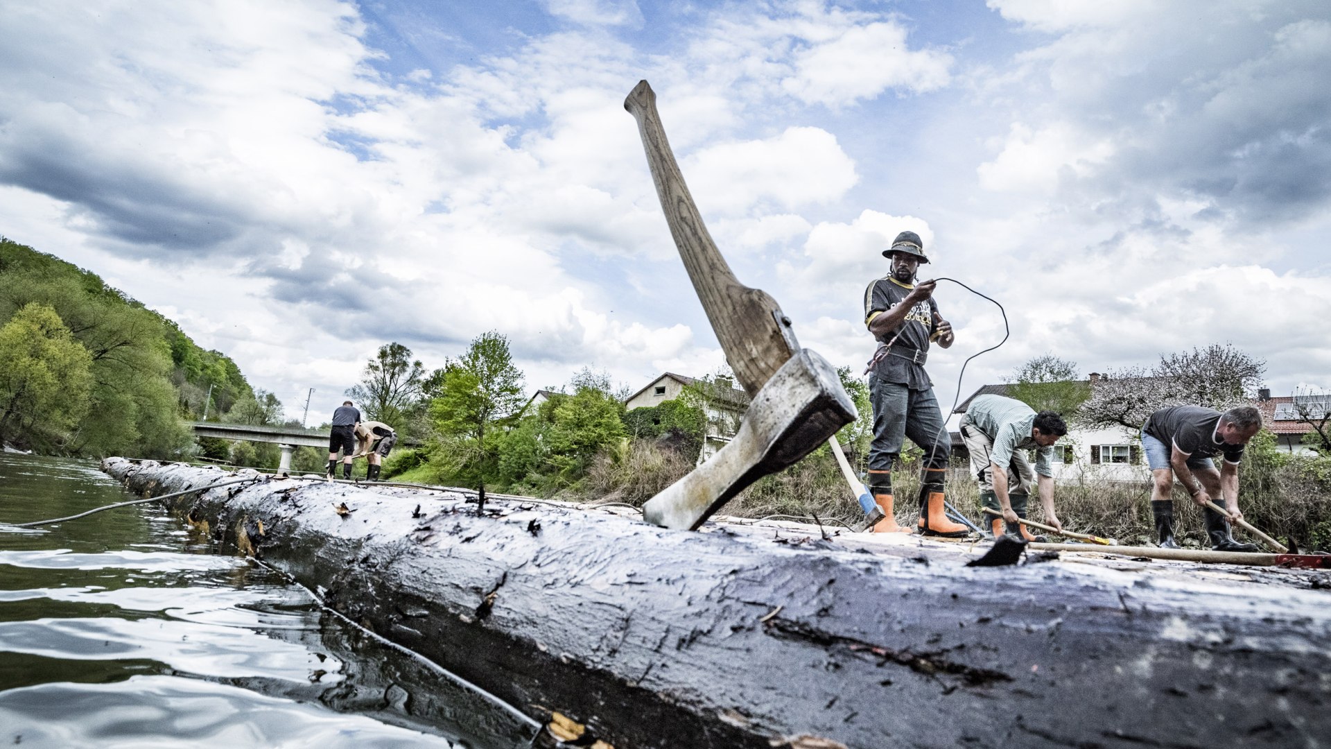 Im Vordergrund steckt eine Axt im Baumstamm eines Floßes, an dem 5 Männer arbeiten, die im Hintergrund zu sehen sind. Das Floß liegt bereits im Wasser. , © Adrian Greiter Im Vordergrund steckt eine Axt im Baumstamm eines Floßes, an dem 5 Männer arbeiten, die im Hintergrund zu sehen sind. Das Floß liegt bereits im Wasser. , © Adrian Greiter