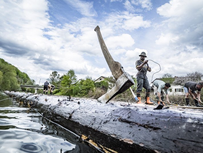 Im Vordergrund steckt eine Axt im Baumstamm eines Floßes, an dem 5 Männer arbeiten, die im Hintergrund zu sehen sind. Das Floß liegt bereits im Wasser. , © Adrian Greiter Im Vordergrund steckt eine Axt im Baumstamm eines Floßes, an dem 5 Männer arbeiten, die im Hintergrund zu sehen sind. Das Floß liegt bereits im Wasser. , © Adrian Greiter