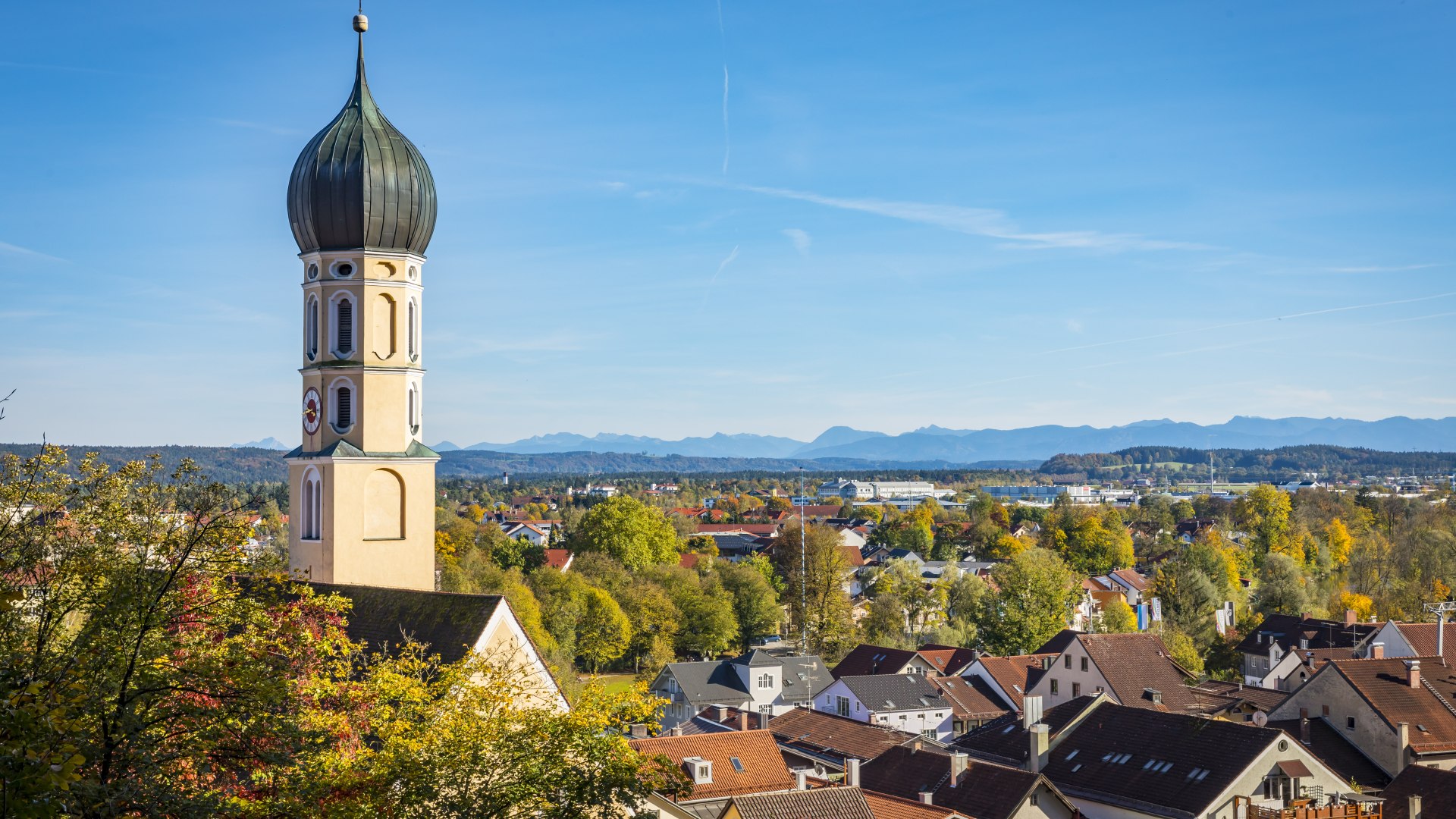 Es ist der Kirchturm und die Dächer von Wolfratshausen bei Sonnenschein zu sehen. Im Hintergrund sieht man weit entfernt die Silhouette der Alpen im Tölzer Land., © Stadt Wolfratshausen, Foto: Adrian Greiter