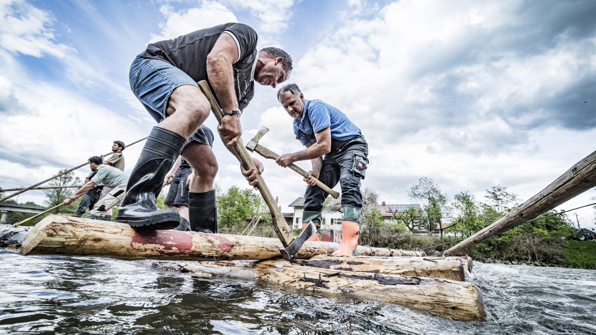Zwei M&auml;nner auf Baumst&auml;mmen, die im Wasser treiben und zu einem Flo&szlig; zusammen gebaut werden, &copy; Stadt Wolfratshausen Foto: Adrian Greiter