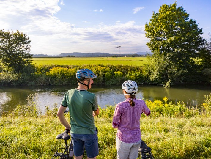 Ein Paar mit Fahrr&auml;dern steht am Isar Loisach Kanal und schaut auf&acute;s Wasser. , &copy; Stadt Wolfratshausen Foto: Adrian Greiter