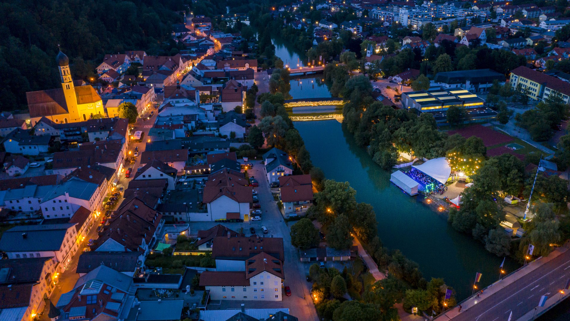 Eine Luftaufnahme vom beleuchteten Wolfratshausen. In der Mitte die Loisach mit dem beleuchteten Sebastiani Steg und dem Festgelände des Fluss Festivals. Das Bild ist aus großer Höhe aufgenommen. , © Adrian Greiter
