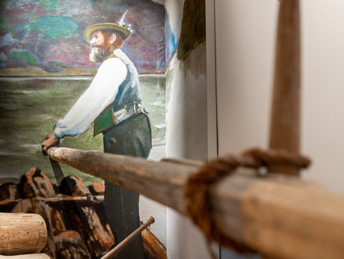 Das Bild zeigt eine Malerei eines Flößers an der Wand, der ein Ruder eines Floßes in der Hand hält, das sich direkt im Raum befindet. , © Stadt Wolfratshausen, Adrian Greiter