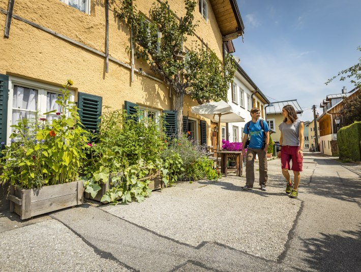 Ein Paar im Wanderoutfit geht an einem gelben Haus am Oberen Obermarkt von Wolfratshausen bei Sonnenschein spazieren., &copy; Adrian Greiter