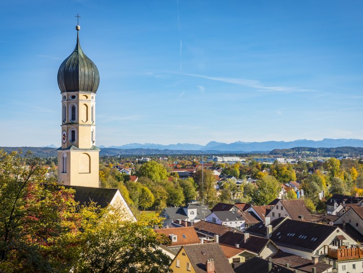Es ist der Kirchturm und die Dächer von Wolfratshausen bei Sonnenschein zu sehen. Im Hintergrund sieht man weit entfernt die Silhouette der Alpen im Tölzer Land., © Stadt Wolfratshausen, Foto: Adrian Greiter