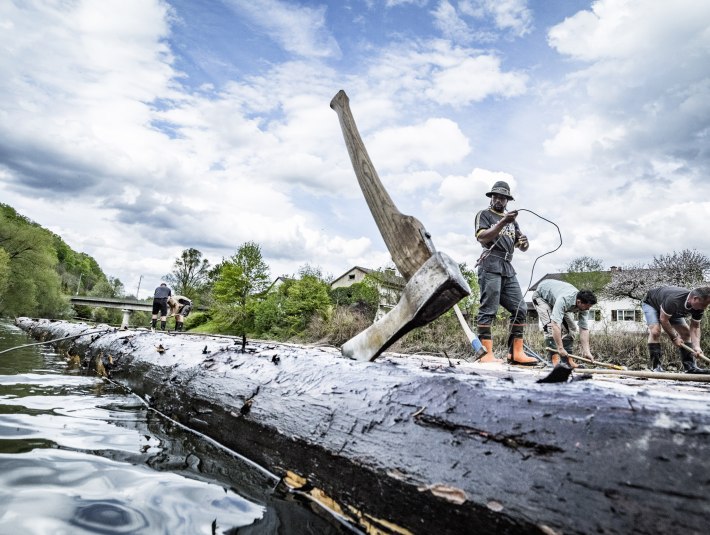 Im Vordergrund steckt eine Axt im Baumstamm eines Floßes, an dem 5 Männer arbeiten, die im Hintergrund zu sehen sind. Das Floß liegt bereits im Wasser. , © Adrian Greiter