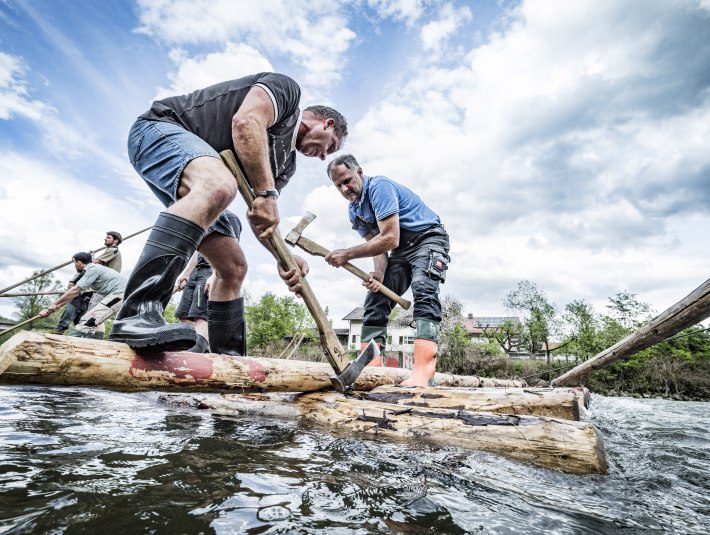 Zwei M&auml;nner auf Baumst&auml;mmen, die im Wasser treiben und zu einem Flo&szlig; zusammen gebaut werden, &copy; Stadt Wolfratshausen Foto: Adrian Greiter